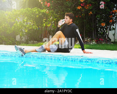 Un uomo dopo un allenamento bevande acqua da un vetro mentre è seduto vicino a una piscina. Foto Stock