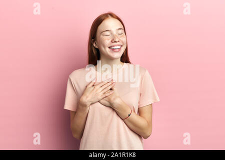 Amichevole donna positivo sorrisi sinceramente, mantiene entrambe le palme sul cuore, vestito in rosa informale di t-shirt, isolato sulla parete rosa, esprime buona attitudine Foto Stock