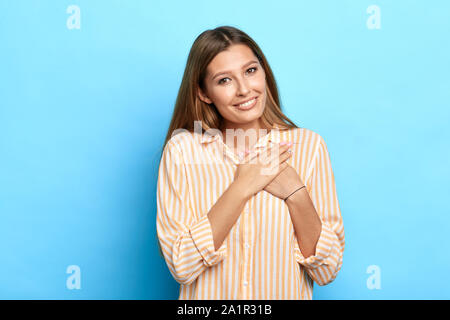 Bella ragazza mantiene entrambe le palme sul cuore, sente gratitudine, essere toccati dalla serenata, vestito di casual camicia a righe, isolate su blue bac Foto Stock