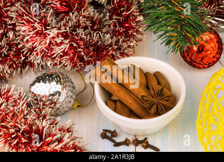 Composizione di natale - bastoncini di cannella, anice stellato e le mandorle in una ciotola bianco, i chiodi di garofano su un tavolo di legno, intorno a loro è rosso tinsel, albero di Natale Foto Stock