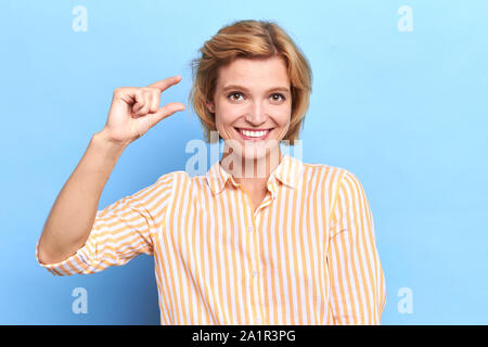 Felice positivo bella donna bionda mostra qualcosa di piccolo con le mani, isolata contro sfondo blu. Bella ragazza dimostra la dimensione di tallone, Foto Stock