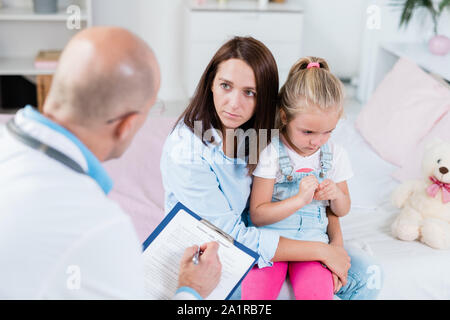 Preoccupato per il giovane madre ascolto ai consigli del medico mentre si tiene il suo malati piccola figlia sulle ginocchia in ospedale Foto Stock