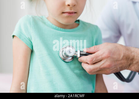 La mano del giovane pediatra tenendo uno stetoscopio mentre mettendolo al torace del malato bambina durante il trattamento medico Foto Stock