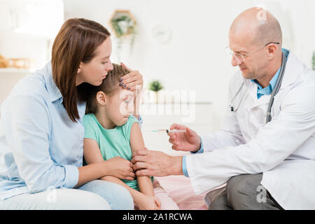 Pediatra contemporanea rendendo iniezione per la piccola ragazza seduta accanto a sua madre in ufficio medico Foto Stock