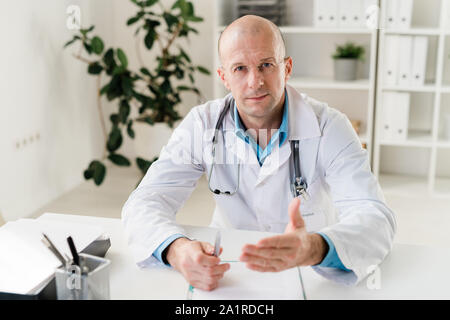 Fiducioso medico con stetoscopio e penna guardando a voi offrendo consulenza medica in ospedale Foto Stock