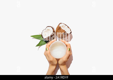 Vista superiore mani femminili vaso di contenimento di naturale di olio di noce di cocco accanto alle noci di cocco fresco e foglie verdi su sfondo bianco, piatto laici. Foto Stock