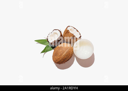 Noci di cocco naturale accanto al vasetto di olio e foglie verdi su sfondo bianco, minima piana composizione laici. Foto Stock