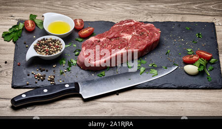 Entrecote o bistecca di ribeye crudo con coltello da cucina, ingredienti freschi e condimenti su un tagliere e sfondo di legno Foto Stock