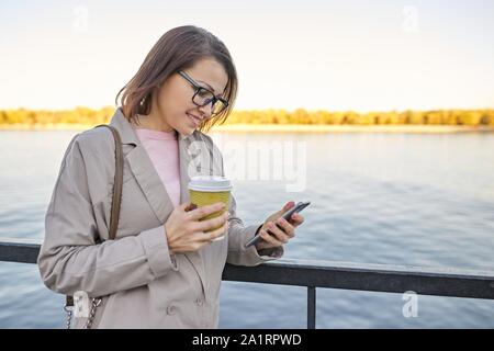Outdoor 40s ritratto di donna, sorridente femmina con tazza di caffè e lo smartphone. Foto Stock