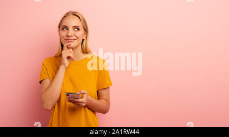 Bionda ragazza carina tipi un messaggio con il suo smartphone. Malinconici ed espressioni di sorpresa di fronte. Sfondo rosa. Spazio vuoto per il testo Foto Stock