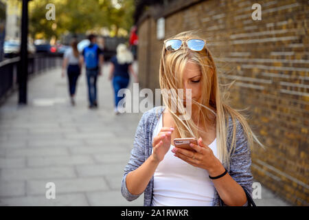 Giovane donna bionda cercando di trovare il modo per il suo telefono per le strade di Londra, Regno Unito, mentre il vento che soffia in suoi capelli Foto Stock