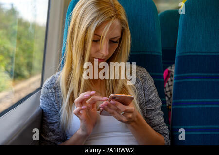 Attraente urbano giovane donna seduta su un treno e controllando il suo telefono cellulare mentre si è in viaggio per il centro di Londra, Regno Unito Foto Stock