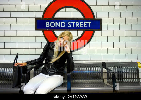 Attraente urbano giovane donna seduta su una panchina di piattaforma a Bond Street e in attesa di una metropolitana di Londra, Regno Unito Foto Stock