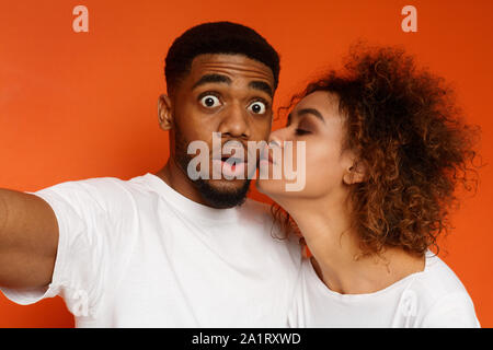 Scioccato ragazzo africano tenendo selfie, donna baciando la guancia Foto Stock