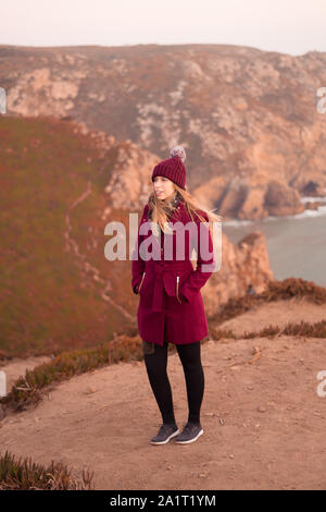 Giovane donna su un cappotto rosso contemplando il mare dalla cima di una scogliera Foto Stock