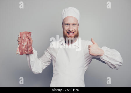 Ritratto di lui attraente bello allegro macellaio positivo guy holding carni fresche nella mano del braccio che mostra il pollice-up isolate su grigio Sfondo pastello Foto Stock