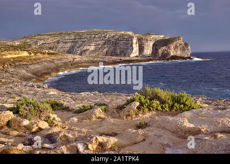 Bella round bay, piante verdi e Fungus Rock Cliffs al tramonto a Gozo. Arancione della luce al tramonto con il grigio nuvole temporalesche. Foto Stock