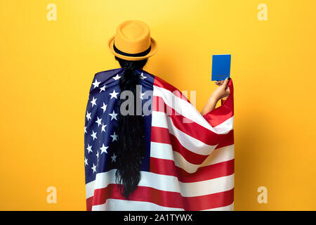 Ragazza sta con la schiena e tiene la bandiera americana su sfondo giallo, ragazza nel quartiere alla moda di hat con bandiera degli Stati Uniti, donna tenendo il passaporto in mano, ho Foto Stock