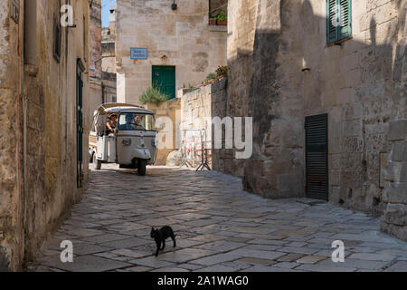 Matera, Italia - 9 Settembre 2019: "Piaggio Ape' 3-wheeler taxi su Via Fiorentini nel cuore dei Sassi di Matera e un gatto che attraversa la strada in pietra Foto Stock