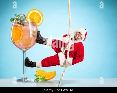 Felice Natale Santa Claus arrampicata su corda vicino da cocktail su sfondo blu. Caucasica modello maschile in costume tradizionale. Concetto di vacanze, Capodanno, umore invernale, doni. Copyspace. Foto Stock