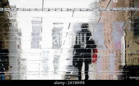Sfocate silhouette di riflessione sul bagnato via della città di una persona che cammina e trasportare valigie di viaggio nella piovosa giornata d'autunno