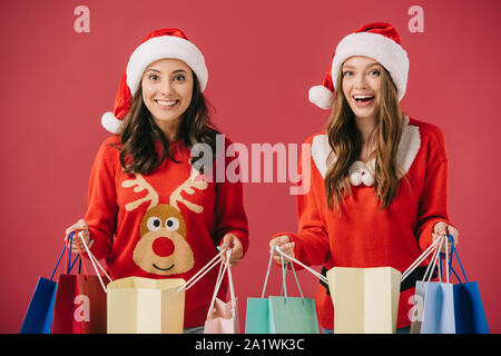 Attraente e sorridente donne in maglioni e cappelli di Babbo Natale azienda shopping bags isolato su rosso Foto Stock