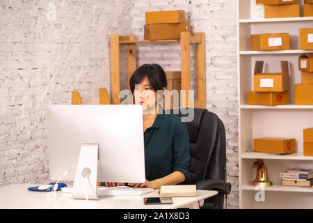 Determinato Asian lady digitando sulla tastiera del computer mentre si lavora con i pacchi in ufficio per la consegna Foto Stock