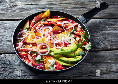 Chilaquiles con prosciutto, sbriciolato panela formaggio, fette di avocado e salsa di pomodoro in una padella su una tavola in legno rustico, cucina messicana, vista orizzontale Foto Stock