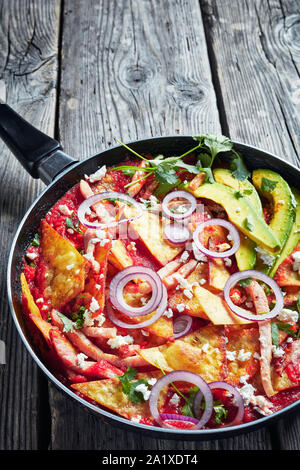 Chilaquiles con prosciutto, sbriciolato panela formaggio, fette di avocado e salsa di pomodoro in una padella su una tavola in legno rustico, cucina messicana, vista verticale fr Foto Stock