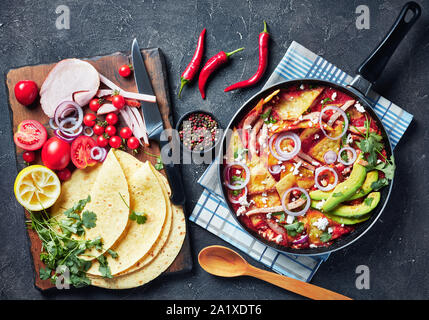 Chilaquiles con prosciutto, sbriciolato panela formaggio, fette di avocado e salsa di pomodoro in una padella su una tabella di calcestruzzo con ingredienti su un tagliere, Me Foto Stock