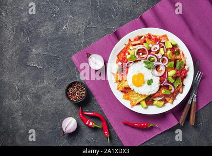 Chilaquiles con un fritto sunny side up di uova, prosciutto, sbriciolato panela formaggio, avocado e salsa di pomodoro su una piastra su un tavolo in cemento, piatto messicano, visualizzare f Foto Stock