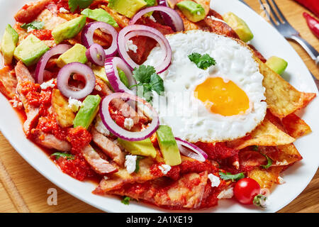 Close-up di chilaquiles fatti in casa con un uovo fritto, prosciutto, sbriciolato panela formaggio, avocado e salsa di pomodoro su una piastra bianca, piatto messicano, vista da abo Foto Stock