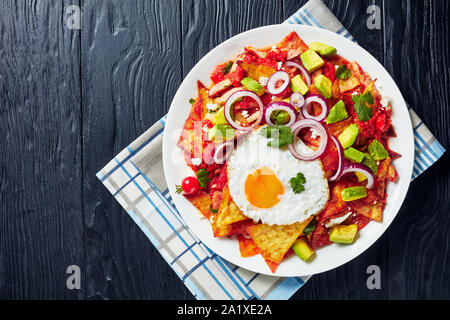 Piccante e speziato colazione messicana di nachos con un uovo fritto, prosciutto, sbriciolato panela formaggio, avocado e salsa di pomodoro su una piastra bianca su di un legno nero t Foto Stock