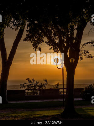 Il sole che illumina un lampione in alberi di un parco affacciato sul mare Foto Stock