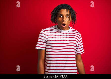 Giovani afro uomo con dreadlocks a strisce che indossa la t-shirt in piedi isolato su sfondo rosso paura e scioccato con sorpresa e stupita espressione, f Foto Stock