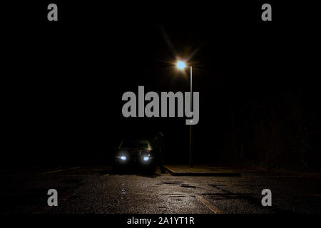 Un spooky, misteriosa figura con cappuccio, in piedi accanto ad un auto sotto una luce di strada di notte Foto Stock