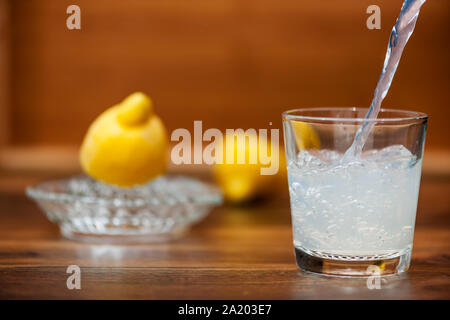 Versando una spremuta di succo di limone in un bicchiere su un tavolo da cucina con piastra dimezzato i limoni e spremitore di limone in sfondo sfocato. Foto Stock