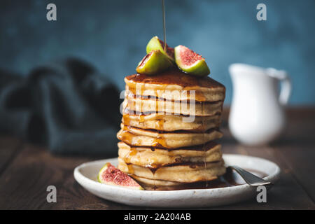 Sciroppo di acero versando sulla pila di frittelle servite con fichi freschi. Dark moody autunno comfort food still life Foto Stock