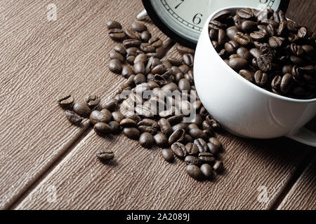 Un orologio con sveglia, i chicchi di caffè in una tazza bianca su di un tavolo di legno. International Coffee Day. Messa a fuoco selettiva. Foto Stock