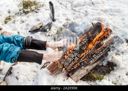 Un uomo si riscalda i suoi piedi scalzi vicino al fuoco nella foresta di pini coperti di neve in inverno Foto Stock