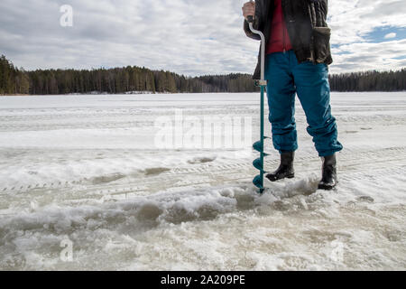 Pescatore di ghiaccio trapani utilizzando un alesaggio sul lago in inverno in una giornata di sole nel bosco Foto Stock