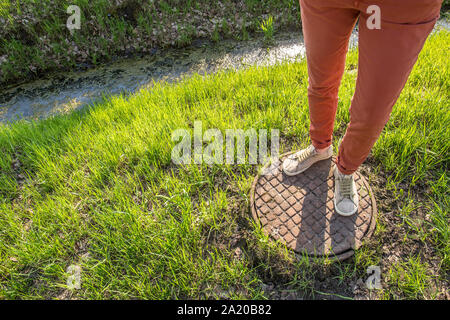 Una persona in piedi su di un vecchio arrugginito chiusino vicino a canaletta con acqua e alghe tra fresco di erba di primavera nel parco Foto Stock