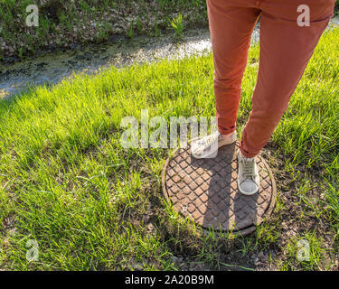 Una persona in piedi su di un vecchio arrugginito chiusino vicino a canaletta con acqua e alghe tra fresco di erba di primavera nel parco Foto Stock