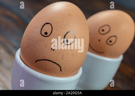 Emozioni concetto due uova sode in bianco d'uovo con coppe disegnate felici e tristi si affaccia su di un tavolo di legno Foto Stock