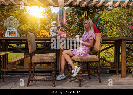 Giovane bella donna bionda in un abito rosa si siede con una tazza di caffè nelle sue mani in un bar o un ristorante in stile vintage di fronte a un vuoto chai Foto Stock