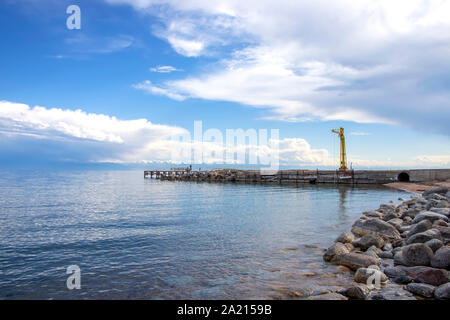 Vista di Issyk-Kul lago, una catena montuosa con cime innevate e un molo da una spiaggia rocciosa. Kirghizistan Foto Stock