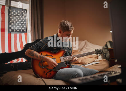 Giovane musicista maschio seduta sul letto in camera da letto gode di trascorrere il tempo a suonare la chitarra a casa Foto Stock