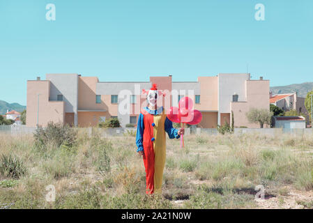 Un inquietante clown indossa un colorato di giallo, rosso e blu all'aperto in costume, tenendo in mano un grappolo di palloncini rossi in mano Foto Stock