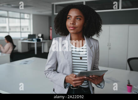 Previsto ben vestito donna giovane azienda digitale compressa perso in pensieri e idee stando in piedi vicino al suo posto di lavoro Foto Stock