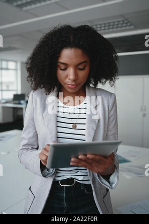Un attraente african american imprenditrice in piedi nella parte anteriore della scrivania utilizzando una tavoletta digitale in un ufficio moderno Foto Stock